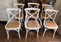 Six white painted wood cross-back dining chairs with woven seats arranged on hardwood floor, front view