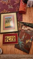 Group arrangement showing four art pieces on floor: Hindu landscape painting in green frame at top left, gold relief in gold frame with red mat at bottom left, large red-brown lacquer panel at right, plus a second wood board.