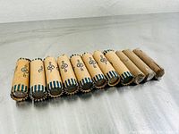 Full view of eleven paper coin rolls, mostly with blue and white striped edges, arranged horizontally on a metallic table; rolls appear sealed and contain old Canadian 5 cent coins.