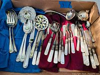 Overview of entire flatware collection including silverplated and mother of pearl handled pieces in blue and red cloth wraps inside a cardboard box.