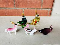 Five miniature animal figurines displayed on a white surface against a brick and siding background: two colorful art glass birds, one dark glass mouse, and two porcelain animals resembling a dog and a rabbit.