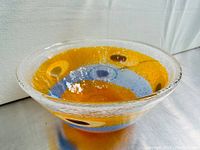 Three-quarter angle view of bowl showing orange center, light blue band, clear textured rim, and oval murrine motifs