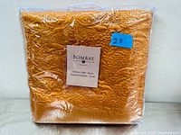 Front view of the orange quilt folded and sealed in clear plastic bag with label.