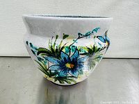 Photo of the front of the white ceramic planter pot showing colorful blue floral and green foliage hand-painted design.