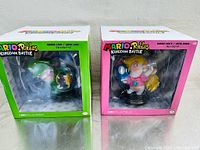 Front view of two boxed action figures, Rabbid Luigi in green box and Rabbid Peach in pink box
