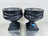 Front view of two black painted concrete urns side by side, showing Grecian style design with swag and fluted details, set on square bases.