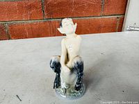 Front view of the Royal Copenhagen porcelain faun figurine on a tree stub against a brick wall background.