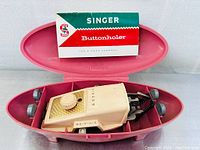 Open pink clam shell case showing beige Singer Buttonholer sewing attachment, instruction booklet inside the lid, 5 metal buttonhole templates placed beside, and a metal bottom plate.