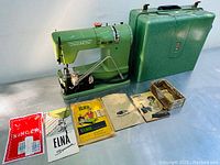 Green vintage Elna Supermatic sewing machine, green hard plastic carrying case, and multiple instruction manuals arranged on a table, showing the lot content and condition.
