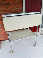 Side view of the vintage gray speckled Formica drop leaf bar cart showing chrome legs and brass wheels