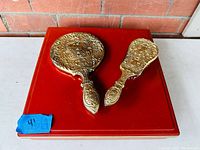Vintage ornate dresser set including a hand mirror and hairbrush with raised floral designs in gold tone resting on a red velvet base