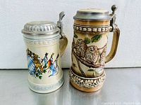 Two vintage porcelain beer steins side by side, one with Bavarian coat of arms design, the other with owl motifs and pewter lids.