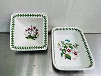 Both the square mini bowl and rectangular trinket tray placed side by side on a metal surface showing their full shape and botanical illustrations.