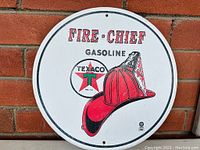 Photo showing full round tin sign with Fire Chief branding, firefighter helmet and Texaco logo against a white background mounted on brick wall.