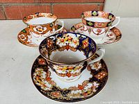 Three vintage porcelain tea cups and saucers with distinct Imari style patterns and colors grouped together on a flat surface.