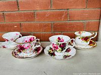 Four Royal Albert fine bone china tea cups with matching saucers featuring various rose patterns, displayed on a grey surface with a red brick wall background.