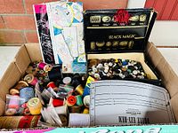 Photo showing a box filled with assorted sewing items including buttons in a Black Magic box, thread spools, and sewing patterns.