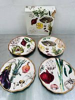 Set of four Williams Sonoma French Market salad plates with rustic scalloped edges, each showing different hand-painted vegetable and herb illustrations, displayed with original box in the background.