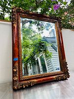 Front view of large ornate mirror with detailed wooden frame and floral carvings showing marquetry inlays, reflecting outdoor scene.