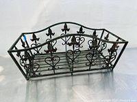Front and side views of rectangular wrought iron flower basket showing heart and fleur-de-lis detailing and open slatted base, aged black finish with some rust spots.