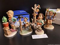 Five M.I. Hummel figurines including Globe Trotter, Apple Tree Boy, Mischief Maker (with original box), Culprits, and Confidentially arranged with a white protective Goebel styrofoam box behind.