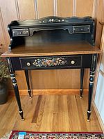 Front view of the entire secretary desk showing the black lacquer finish, brown wooden top, and floral stencil details on drawers and legs.