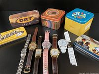 All six Fossil watches arranged next to five colorful empty Fossil tins on black background, showing watch face and band condition plus tin designs.
