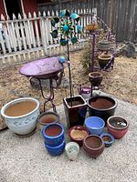 Overall view of purple metal plant stand, bird bath on stand, whirligig spinner, and grouping of assorted garden pots on concrete.