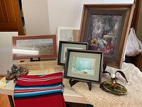 Photo shows assortment of Western art and decor on a table including the Radcliffe Pewter statue, red woven tapestry, framed prints, and the larger framed painting on stand