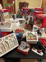 Wide view of multiple boxed and unboxed Santa's Workbench porcelain Christmas village buildings, figurines, accessories, and boxed lights displayed on a table