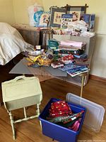 Wide shot showing fabric, patterns, sewing tools on table, and painted mahogany sewing cabinet on legs below table.