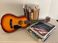 Full view of the sunburst First Act Discovery FG-130 acoustic guitar with several music books behind and beside it.