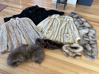 View showing all 3 different women's short fur coats laid flat on wooden floor. Shades include black, cream/light beige, and greyish. Also four fur collars arranged in front of coats.