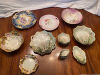 Photo of 10 antique plates and bowls arranged on wooden surface, showing assorted floral china pieces with blue, pink, and lily of the valley themes.