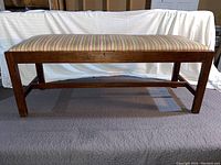 Front view of wooden bench with striped upholstered seat resting on gray surface with white cloth backdrop.