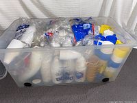 Full view of the large clear plastic bin containing disposable cups, plastic holders in blue, white, and yellow, and other items. Bagged disposable cups labeled Dixie are visible inside.