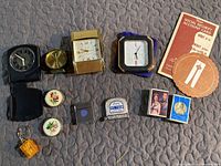 Overview of entire lot showing several travel clocks, measuring tapes, keychain with mini cards, matchboxes, and paper coasters.