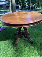 Round wood pedestal table outdoors on grass with visible carved legs and base details.