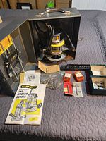 Photo showing Stanley router inside metal case with power cord, two side handles, adjustable depth collar, several boxed bits, instruction manual, and additional accessories laid out