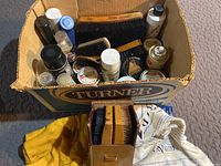 Overview of box filled with various shoe polishing brushes and supplies along with a brown leather carrying case containing brushes and a cloth bag