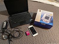 Photo showing black netbook laptop, power adapter, pink iPod Nano, white iPhone with cracked screen, and boxed desk phone with LCD touch panel on a fabric surface.