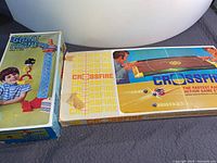 Photo of two vintage game boxes side by side: Giant Crane toy box and Crossfire game box showing box condition and graphics.
