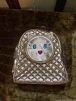 Front view of gold quilted backpack with cat face design featuring crystal eyes, heart nose, and whiskers.