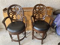 Two wooden barstools with black leather seats and ornate cut-out backrests positioned side-by-side in an indoor garage setting, showing overall appearance and condition.
