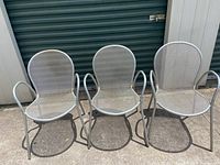 Three silver stackable metal mesh armchairs lined up outside showing entire chairs with armrests and rounded backs.