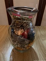Clear glass vase filled with assorted costume jewelry, visible metal chains, beads, faux pearls, and some packaging materials inside.