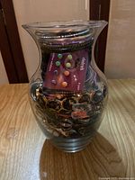 Clear glass vase filled with assorted costume jewelry showing various metal-tone bracelets, a card of pastel-colored stud earrings, and other jewelry pieces.