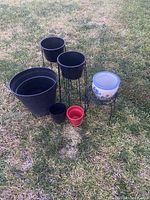 Set of six outdoor flowering pots in various sizes and colors, including two pots on black metal stands placed on grass.