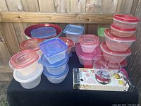 Photo showing various plastic food storage containers in red, blue, and clear lids, two large red mixing bowls in back, small glass measuring cup in front, cutting board underneath glass cup, gripper whisk on top.