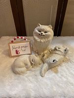 Photo showing all three ceramic cat figurines and the small wall art sign on a soft beige surface with a brown wood background.